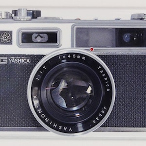 May include: A silver and black 35mm film camera with a black lens. The lens has the text "Yashica" and "Japan" printed on it. The camera has a silver top with a black viewfinder and a silver bottom with the text "Yashica" printed on it.