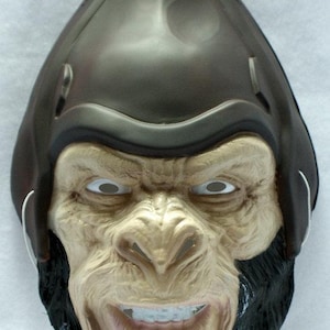 Könnte beinhalten: Eine braune und beige Gorillamaske mit schwarzem Bart und einer dunkelbraunen, helmähnlichen Kopfbedeckung. Die Maske hat weiße Augenöffnungen und einen lächelnden Mund. Die Maske besteht aus Kunststoff und hat eine schwarze Aufhängelasche.