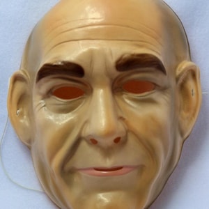 May include: A vintage-style mask depicting a bald man's face. The mask is flesh-toned with brown eyebrows and a pink mouth. It has eye holes and a string attached for wearing. The mask is likely made of plastic or a similar material.