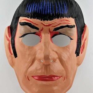 May include: A vintage mask depicting a character with a tan face, black hair with blue highlights, and red accents around the eyes and ears. The mask has eye cutouts and red lips, and is likely made of paper or cardboard.