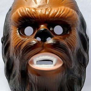 Könnte beinhalten: Eine braune Plastikmaske von Chewbacca mit schwarzen Haaren und weißen Augenlöchern. Die Maske hat eine Mundöffnung mit weißen Zähnen.