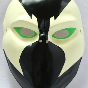 Op de afbeelding: Een wit en zwart masker met groene oogaccenten. Het masker heeft een glanzende afwerking en een opvallend ontwerp, met zwarte vormen op een witte basis. De oogopeningen zijn groen omlijnd, met een neutrale kleur binnenin.