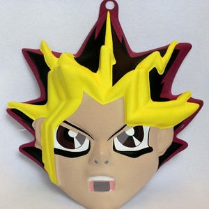 Pode incluir: Um rosto decorativo e estilizado de um personagem com cabelo amarelo espetado, olhos castanhos e pele clara. O rosto do personagem está sobre um fundo em forma de estrela com uma borda vermelha escura e detalhes em preto. Há um pequeno buraco no topo.