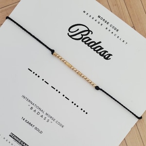 Op de afbeelding: Een zwart koordarmband met gouden kralen die "BADASS" in morsecode spellen. De armband wordt gepresenteerd op een witte kaart met de woorden "MORSE CODE MESSAGE BRACELET" en "14 KARAT GOLD".