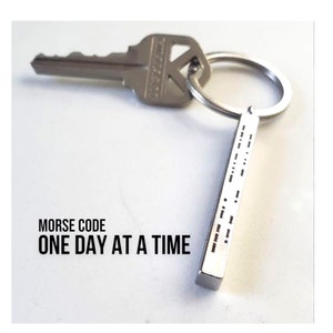 May include: A silver key and a keychain with a Morse code design. The keychain has the text "MORSE CODE ONE DAY AT A TIME". The keychain is rectangular and has a series of dots and dashes.