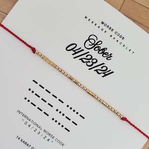 Puede incluir: Un brazalete de código Morse con cuentas doradas sobre un hilo rojo, colocado sobre una tarjeta blanca. La tarjeta tiene las palabras "Sober 04/23/24" y "Morse Code Message Bracelet" impresas. El brazalete está hecho de pequeñas cuentas doradas rectangulares.