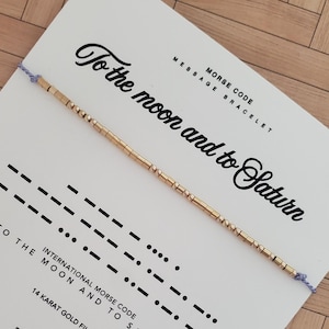 Op de afbeelding: Een goudkleurige Morsecode armband met de boodschap "To the moon and to Saturn" op een witte kaart. De kaart toont ook de internationale Morsecode voor de boodschap. De armband is gemaakt van 14 karaats goud.