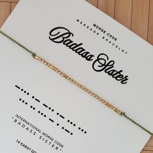 May include: A gold beaded bracelet on a white card with the words "Badass Sister" in a script font. The card also displays the Morse code for "Badass Sister" and the text "Message Bracelet". The bracelet is strung on a green cord.