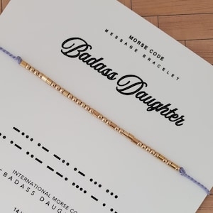 May include: A gold beaded Morse code bracelet on a white card with the words "Badass Daughter". The card also includes Morse code symbols and the text "Morse Code Message Bracelet". The bracelet is strung on a light purple cord.