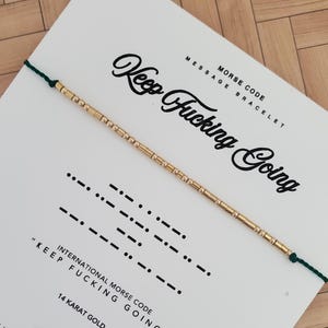 Könnte beinhalten: Ein goldfarbenes Morsecode-Armband mit der Aufschrift "Keep Fucking Going" auf einer weißen Karte. Die Karte zeigt auch die Morsecode-Übersetzung und den Text "14 Karat Gold". Das Armband ist an einem grünen Band befestigt.
