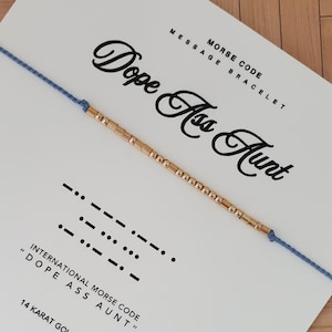 Könnte beinhalten: Ein goldfarbenes Morsecode-Armband an einem hellblauen Band, das "Dope Ass Aunt" buchstabiert. Das Armband wird auf einer weißen Karte mit dem Text "Morse Code Message Bracelet" angezeigt.