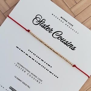 May include: A gold-filled Morse code bracelet on a white card with the words "Sister Cousins". The bracelet is strung on a red cord. The card also includes the text "International Morse Code" and "14 Karat Gold Fill".