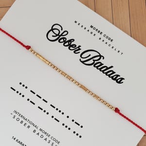 May include: A gold beaded bracelet with a red cord, presented on a white card. The card features the words "Sober Badass" in elegant script, along with Morse code symbols and the text "Morse Code Message Bracelet."