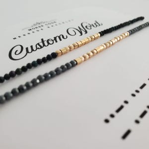 Könnte beinhalten: Zwei Halsketten mit Perlen, goldfarbenen Akzenten und dunkelgrauen Perlen. Die Halsketten liegen auf einer weißen Oberfläche mit den Worten "Custom Word" in eleganter Schrift. Morsecode ist ebenfalls sichtbar.