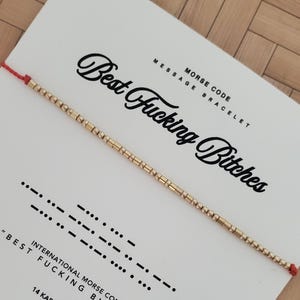 Op de afbeelding: Een goudkleurige morsecode-armband met de tekst "Best Fucking Bitches" op een witte kaart. De kaart bevat ook de morsecodevertaling en de woorden "Morse Code Message Bracelet."