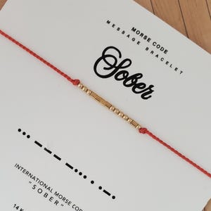 May include: A red string bracelet with gold beads spelling "Sober" in Morse code. The bracelet is displayed on a white card with the words "Morse Code Message Bracelet" and the word "Sober" in cursive.