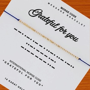 May include: A blue cord bracelet featuring gold-colored beads arranged in Morse code, displayed on a white card printed with the text "MORSE CODE MESSAGE BRACELET", "Grateful for you", and "INTERNATIONAL MORSE CODE "GRATEFUL FOR YOU" 14 KARAT GOLD".