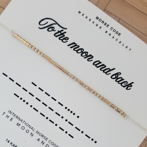 Op de afbeelding: Een goudkleurige Morsecode-armband op een witte kaart met de tekst "To the moon and back". De kaart bevat ook Morsecode-symbolen en de woorden "Morse Code Bracelet".