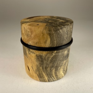 May include: A cylindrical wooden container with a lid. The wood features a natural, swirling grain pattern in shades of tan and grey. A black band encircles the middle of the container, adding a contrasting detail.