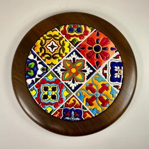 May include: Circular wooden trivet with a colourful ceramic tile insert. The tile design features a vibrant patchwork of floral and geometric patterns in red, yellow, blue, and green. The trivet is designed to protect surfaces from hot items.