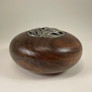 May include: A dark brown wooden bowl with a silver-colored, leaf-like design on the top. The bowl has a smooth, rounded shape and is likely used for decorative purposes.