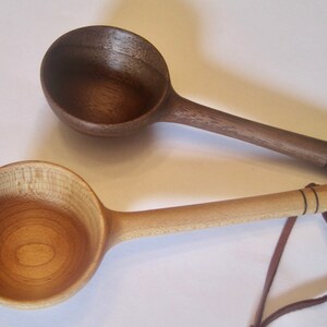 May include: Two wooden spoons with rounded bowls and long handles. One spoon is dark brown, the other light tan. Both have a leather strap attached to the handle's end. The spoons are likely for cooking.