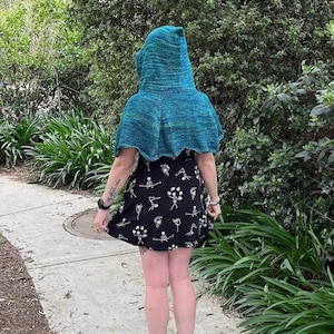 Puede incluir: Una persona lleva una capa con capucha turquesa con un vestido negro con estampado de esqueleto. La persona está de pie en un camino pavimentado con vegetación verde en el fondo.