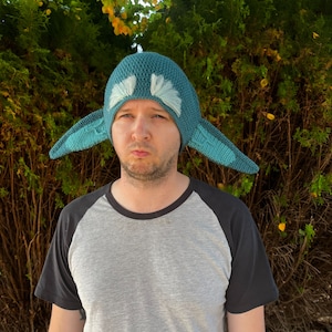 May include: A person wearing a teal blue knitted hat with white fur accents and ear flaps. The hat is shaped like a creature with large ears. The person is wearing a grey and black baseball t-shirt.