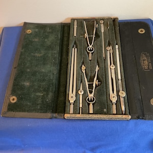 Op de afbeelding: Een open, vintage tekenset in een zwarte koffer met een groene fluwelen binnenkant. De set bevat verschillende zilverkleurige passer, verdelers en andere tekengereedschappen. De koffer is open op een blauw oppervlak.