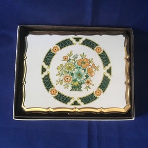 May include: A white metal cigarette case with a gold trim and a floral design. The design features a basket of flowers in shades of orange, yellow, green, and blue, surrounded by a green and gold border.