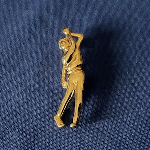 Peut inclure: Épingle en métal doré représentant un golfeur en plein swing. L'épingle montre un golfeur avec le bras levé, tenant un club de golf. L'épingle est posée sur un fond bleu foncé.
