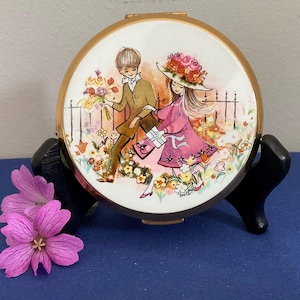 May include: A vintage compact mirror with a gold-tone rim and a painted scene of a couple in a garden. The illustration features a man with flowers and a woman in a pink dress and hat. The compact is displayed on a black stand.