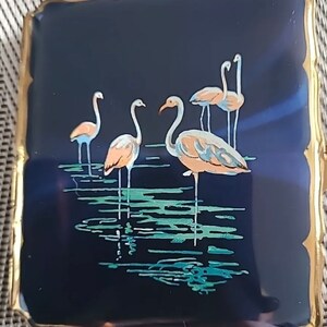 May include: A square, blue and gold compact with a painted design of four pink flamingos standing in a blue and green pond.