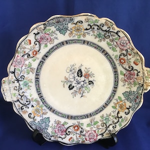 Könnte beinhalten: Eine weiße Keramik-Servierplatte mit gewelltem Rand und Blumenmuster. Die Platte hat einen goldenen Rand und schwarze florale Akzente.