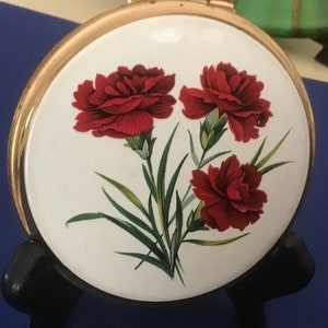 Op de afbeelding: Een ronde, vintage compactpoederdoos met een goudkleurig metalen frame. De witte deksel heeft een bloemenontwerp van drie rode anjers met groene bladeren. De compactpoederdoos wordt gepresenteerd op een zwarte standaard tegen een blauwe achtergrond.