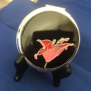May include: A silver compact mirror with a black background and a pink and red lily flower design.