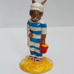 Peut inclure: Une figurine de lapin brun portant un maillot de bain rayé bleu et blanc et un masque de sommeil blanc, tenant un seau rouge avec du sable jaune. Le lapin est debout sur une base jaune avec des empreintes de pas blanches.