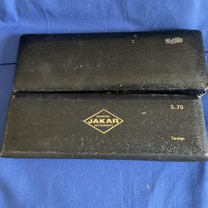 Puede incluir: Dos estuches rectangulares negros, posiblemente para instrumentos de dibujo, apilados sobre una superficie azul. El estuche inferior presenta un logotipo dorado en forma de diamante con el texto "DRAWING JAKAR INSTRUMENTS" y el texto "S.70 Foreign".