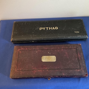 Op de afbeelding: Twee vintage dozen op een blauw oppervlak. De bovenste doos is zwart met het woord "PYTHAG" in gouden letters. De onderste doos is bordeauxrood met een rechthoekig zilveren label. Op de bovenste doos staat ook het woord "Foreign".