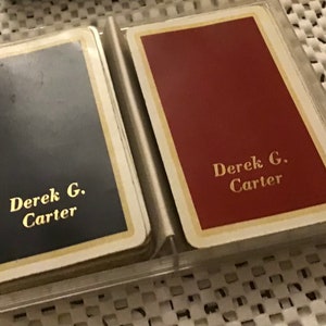 Puede incluir: Dos barajas de cartas de juego con nombres personalizados, "Derek G. Carter", una con el dorso negro y otra con el dorso rojo.