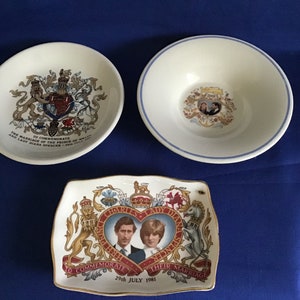 Pode incluir: Três pratos comemorativos. Dois são redondos, um com borda azul e outro com brasão. O terceiro é retangular, apresentando uma imagem em forma de coração de Carlos e Diana, com o texto "To Commemorate Their Marriage 29th July 1981."