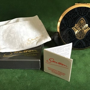 Könnte beinhalten: Ein schwarzer und goldener Kompakt-Spiegel mit einem dekorativen Design. Der Spiegel befindet sich in einer schwarzen Box mit einem weißen Satinbeutel und einer kleinen Karte mit der Aufschrift "Exclusively Designed Handbags & Cosmetic Accessories".