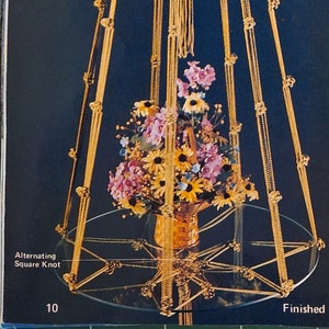 May include: A gold macrame plant hanger with a round top and three legs. The hanger is 40 inches tall and features a variety of knots, including square knots, button knots, and half hitches. The hanger is decorated with a glass vase filled with flowers.