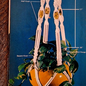 May include: A white macrame plant hanger with a 46-inch finished length. The hanger is made with square knots and half knots. It is holding a terracotta pot with a green plant.