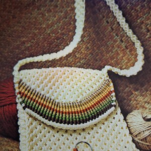 May include: Cream-colored macrame shoulder bag with a flap featuring a rainbow stripe design. The bag has a long, braided strap and a matching keychain. A key hangs from the keychain. The bag is made of a woven material.