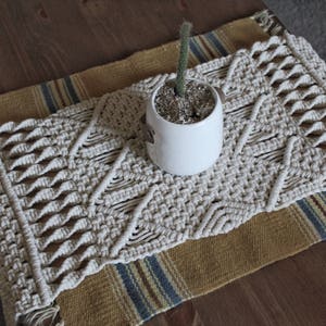 May include: A white macrame table runner with a geometric pattern. The runner is on top of a brown and blue striped rug. A small white planter with a green succulent is on top of the runner.