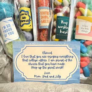 May include: Several clear plastic bags filled with colorful candies and treats. Each bag has a decorative label with phrases like "You're Unstoppable" and "Enjoy the little things." A card reads "I love that you are enjoying everything that college offers."