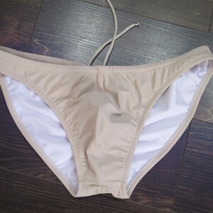 May include: A beige swimsuit bottom with a white lining. The swimsuit has a low rise and a simple design.