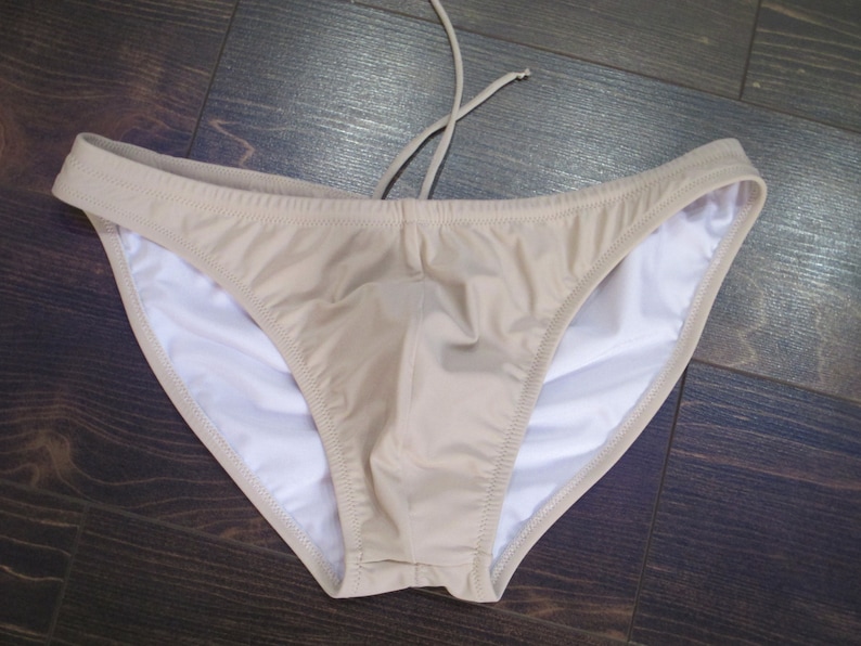 May include: A beige swimsuit bottom with a white lining. The swimsuit has a low rise and a simple design.
