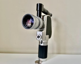 May include: Vintage silver and black movie camera with a large lens. The camera has a black handle and a silver body with a dial and a black grip. The lens reflects the surrounding environment.